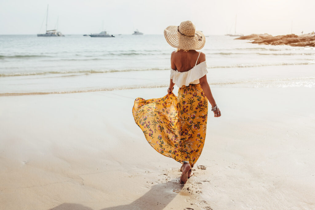 Žena chodící po pláži ve slaměném klobouku a žlutých vzdušných šatech s květinovým vzorem s bílým topem á la halenka - módní šaty na léto 2023