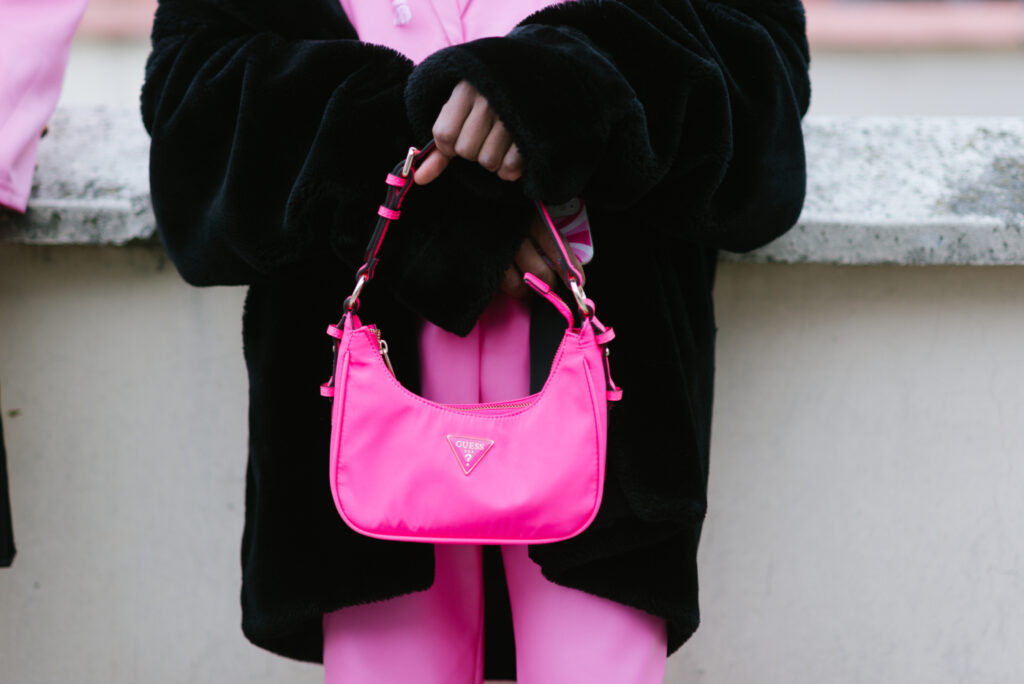Zářivě růžová kabelka Guess v ruce ženy v oversize plisovaném kabátě a růžovém kostýmku