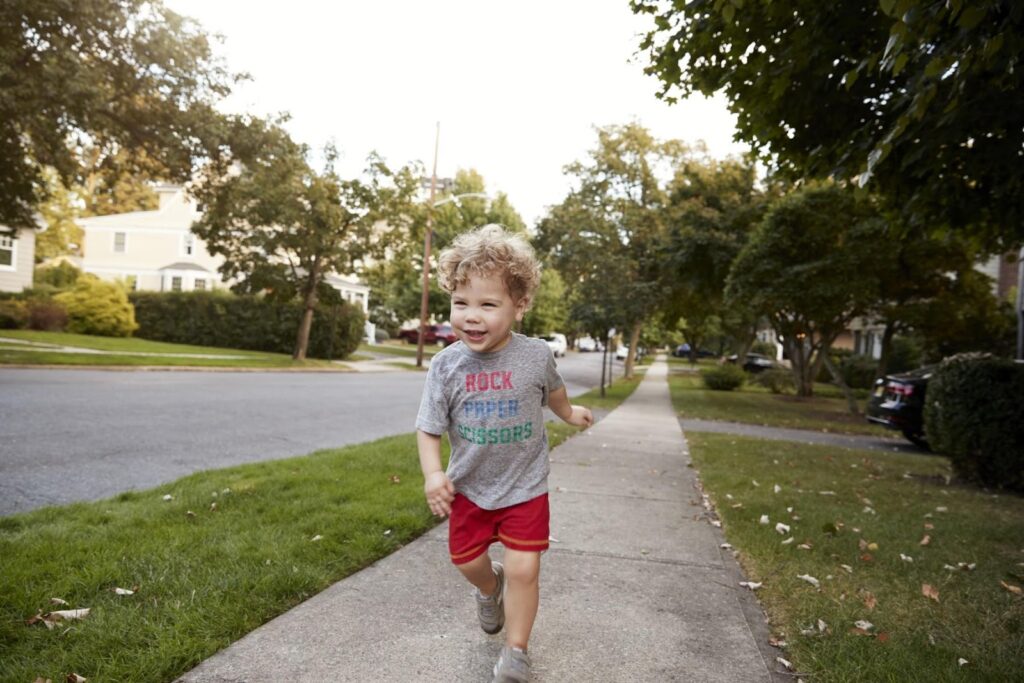 Po chodníku běží malý chlapec v šedém tričku s nápisem a červených šortkách