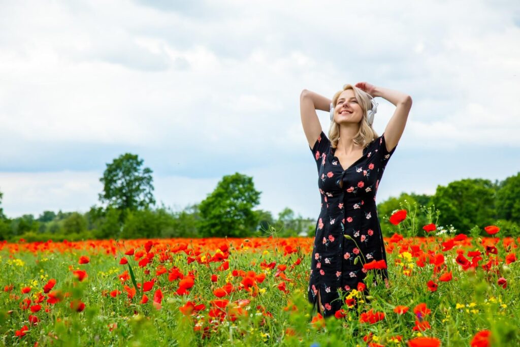 Žena v dlouhých černých šatech s květinami. Stojí v trávě mezi červenými máky. V uších má sluchátka