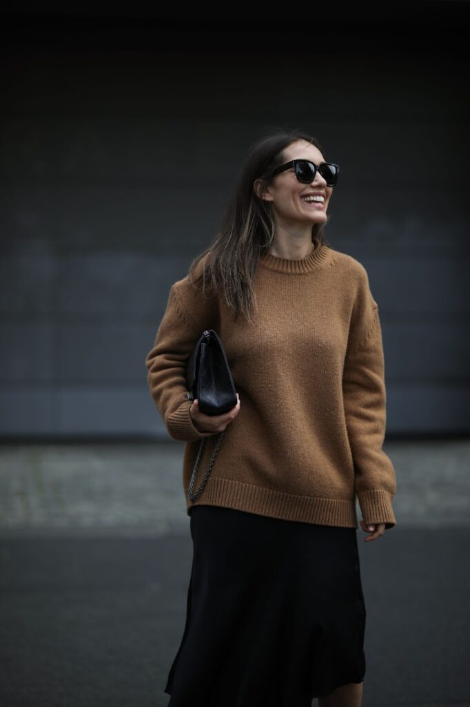 Žena v hnědém svetru v ruce drží černou kabelku. Na obličeji má černé sluneční brýle