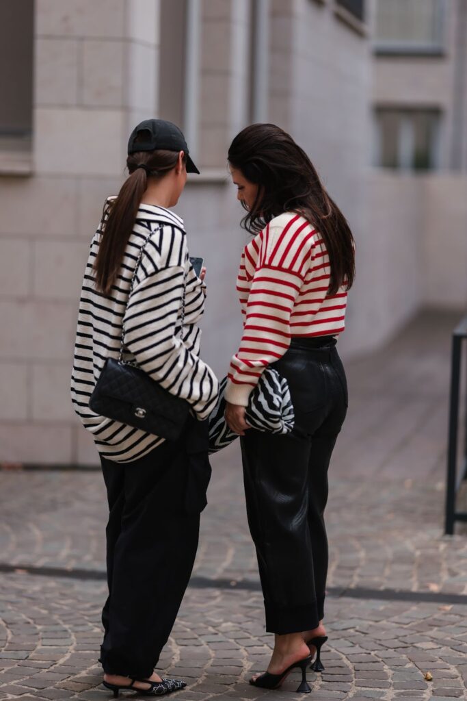 Dvě mladé ženy v pruhovaných tričkách s dlouhými rukávy a černými kalhoty. Dívají se do mobilu