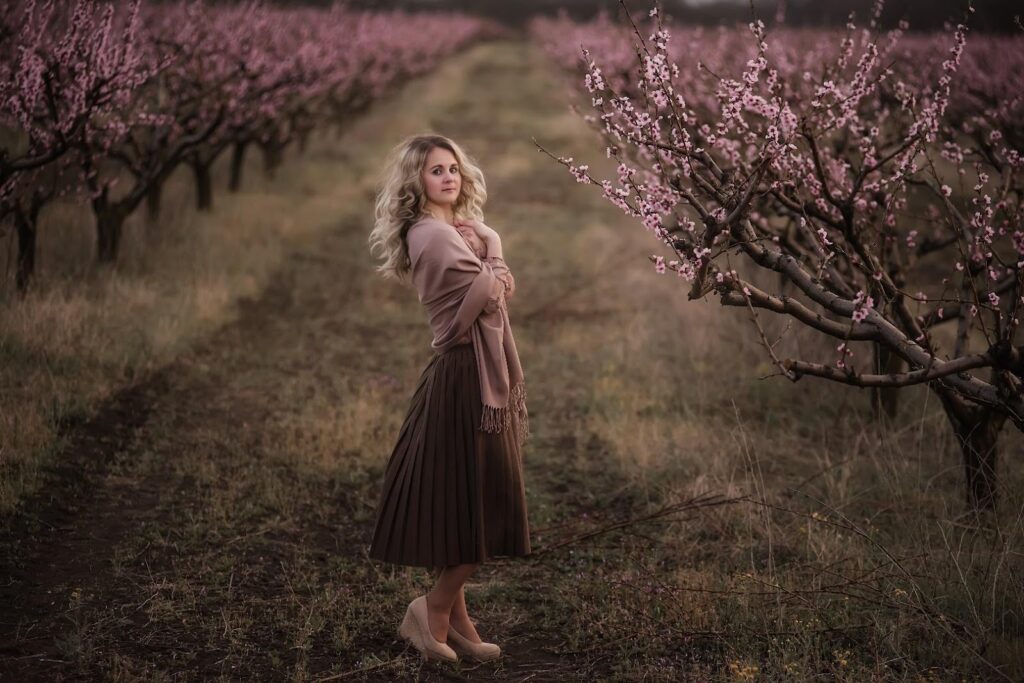 Žena v hnědé plisované sukni stojící v sadu mezi kvetoucími stromy