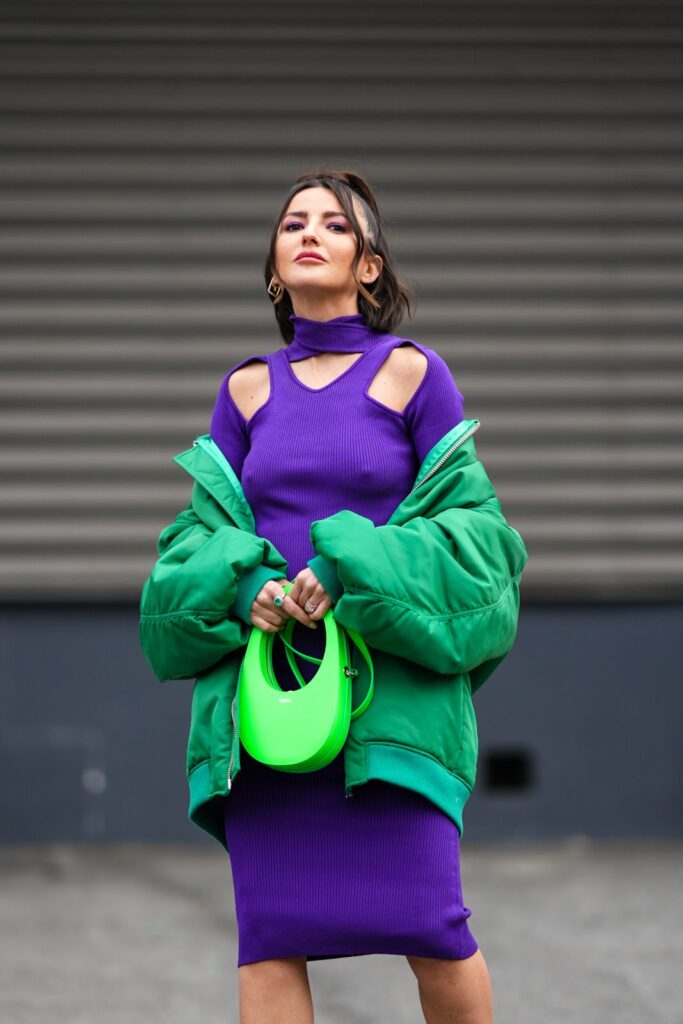 žena ve fialových úpletových šatech, v zelené bomber bundě a se zelenou kabelkou