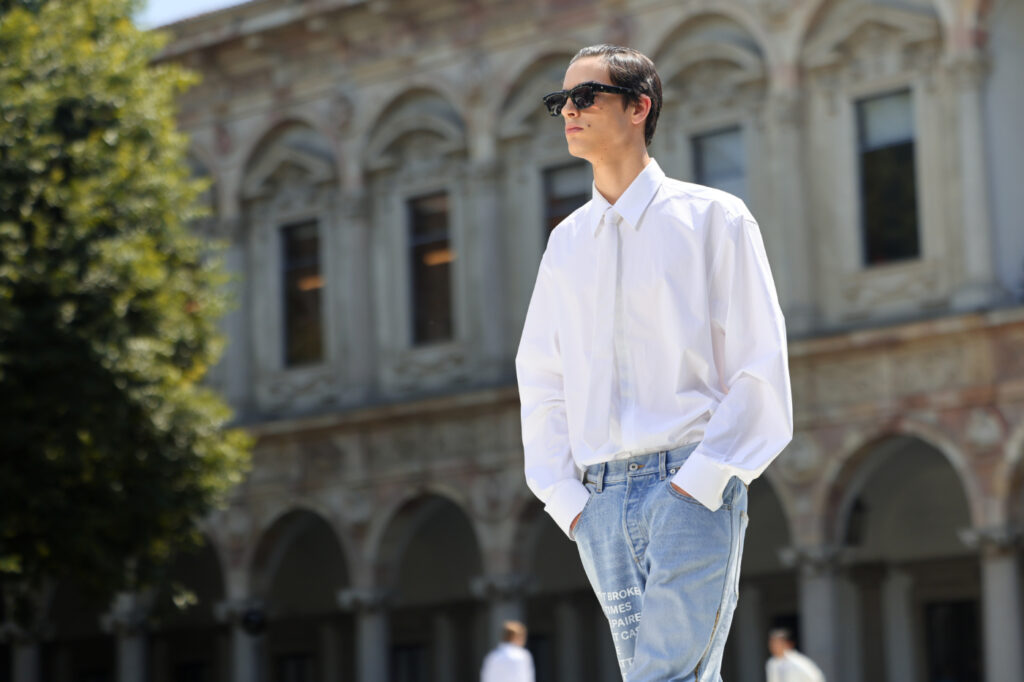 Pánská móda 2024: model ve sněhobílé košili, bílé kravatě, světlých džínách a slunečních brýlích.