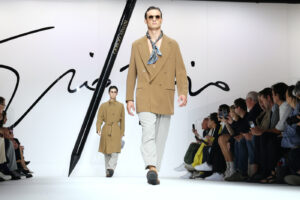 Pánská móda: trendy jaro/léto 2024 na přehlídkových molech – model v kabátu ve velbloudí hnědé a šátkem na krku