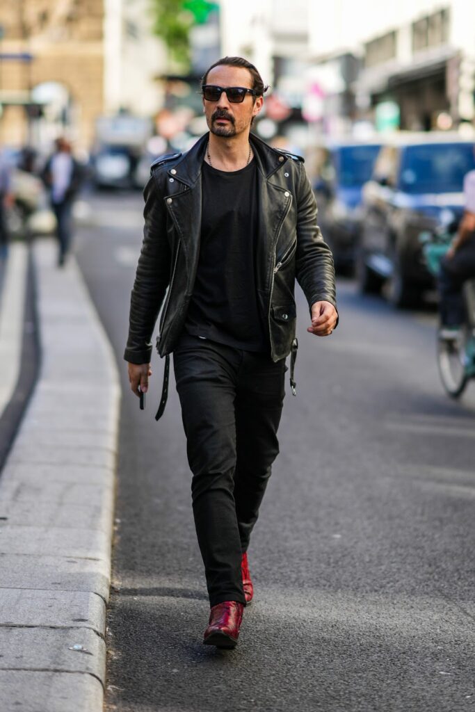 Muž, který jde po ulici. Má na sobě koženou bundu a červené boty. Na nose má sluneční brýle.