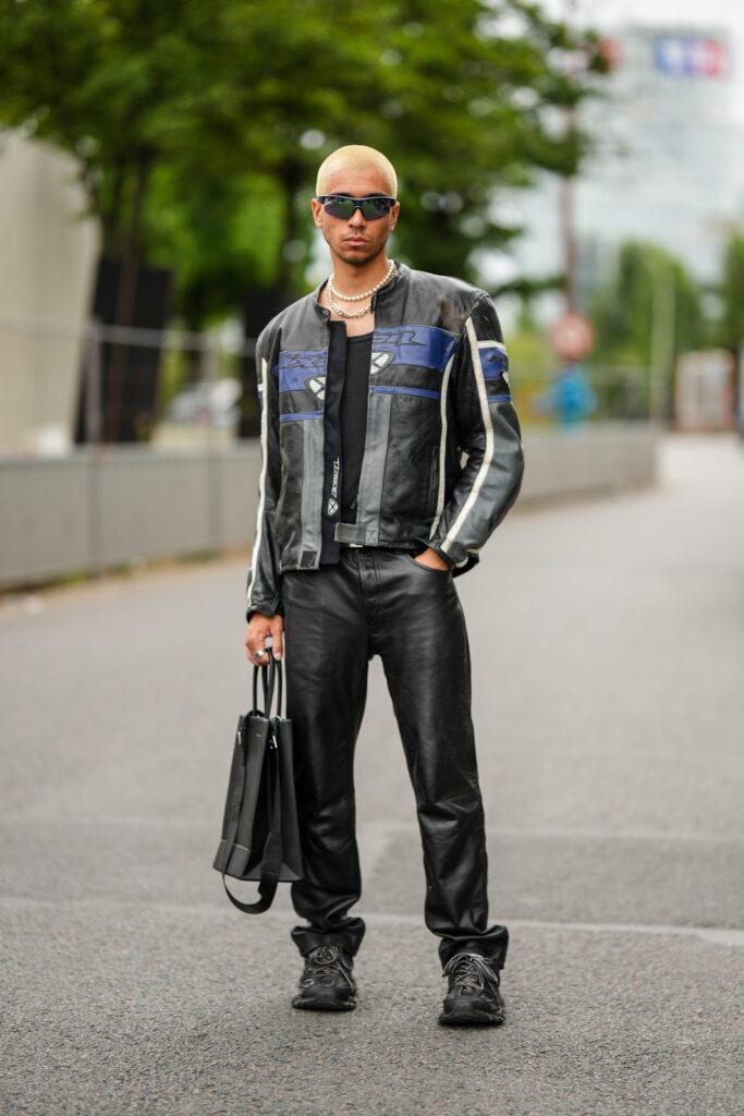 Muž, který stojí na ulici. Má na sobě motorkářskou bundu. V ruce drží tašku a na nose má sluneční brýle.