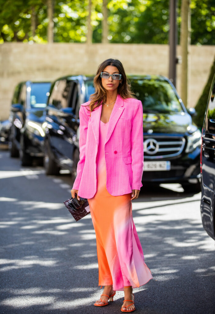 Žena, která stojí na ulici. V pozadí stojí auta. Má na sobě růžové sako a v ruce drží černou kabelku.