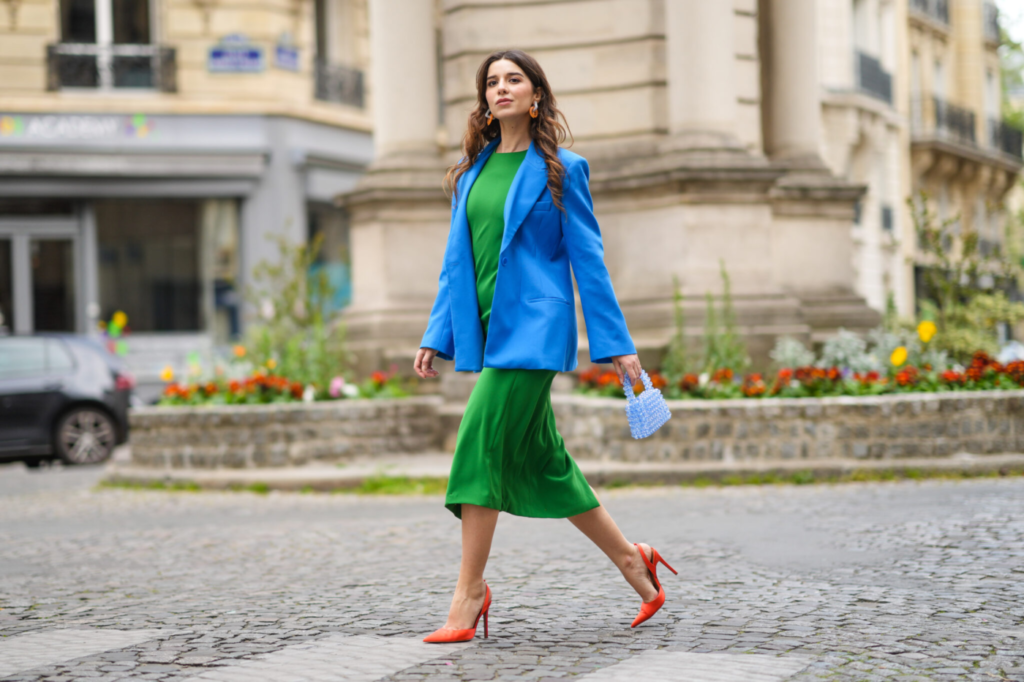 žena v klasických šatech v odstínu svěží zelené, s modrým sakem, modrou kabelkou a červenými jehlovými podpatky.